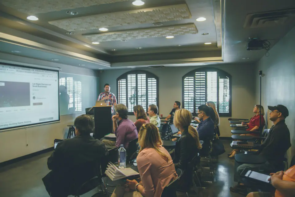 A group of adults seated in rows in a teacher professional development session, taking notes on laptops and in notebooks.